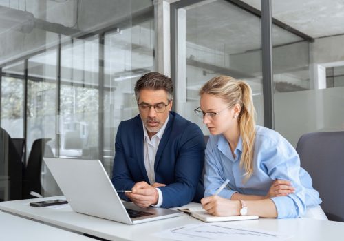 Two partners, team of professional business people working, discussing, writing notes at desk. Mature focused senior mentor leader man showing young woman manager project on laptop in modern office