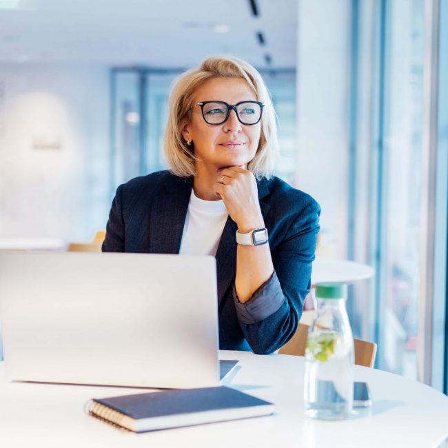 50's confident mature businesswoman dreamy looking at window, middle-aged experienced senior female professional working on laptop in open space office. Female entrepreneur working remotely
